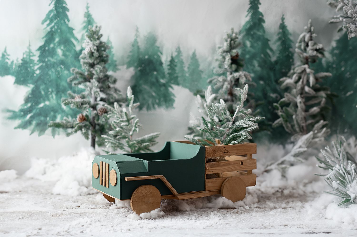 Toy wooden green truck carrying mini Christmas trees in a snowy forest backdrop.