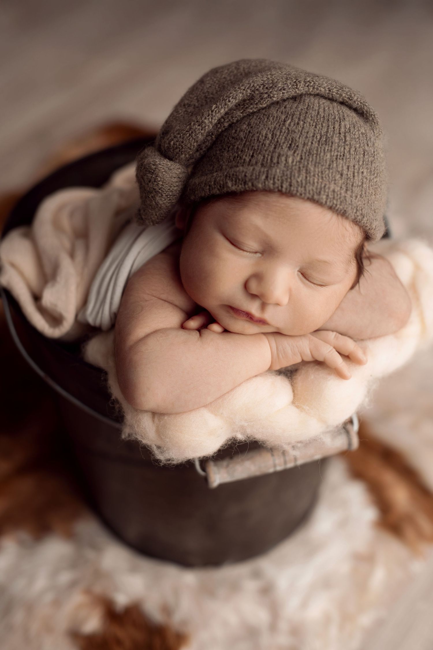 Adorable sleeping newborn in knit hat posed with crossed arms in rustic metal bucket with soft blankets.