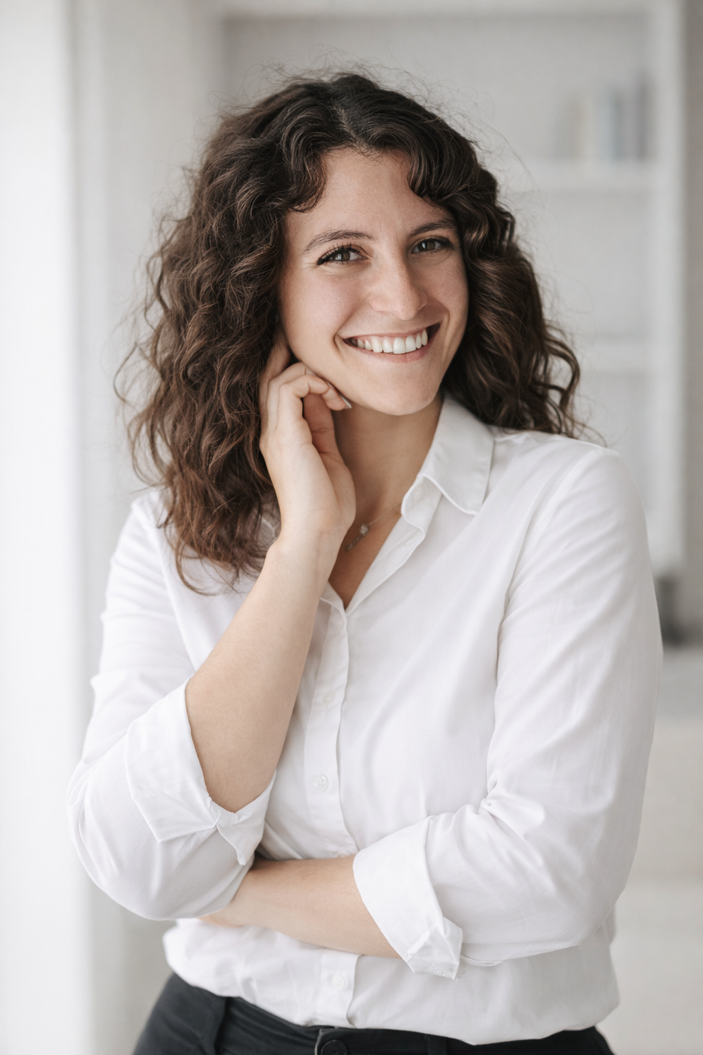 Happy curly-haired woman in white button-up shirt smiling warmly with hand raised to cheek indoors.