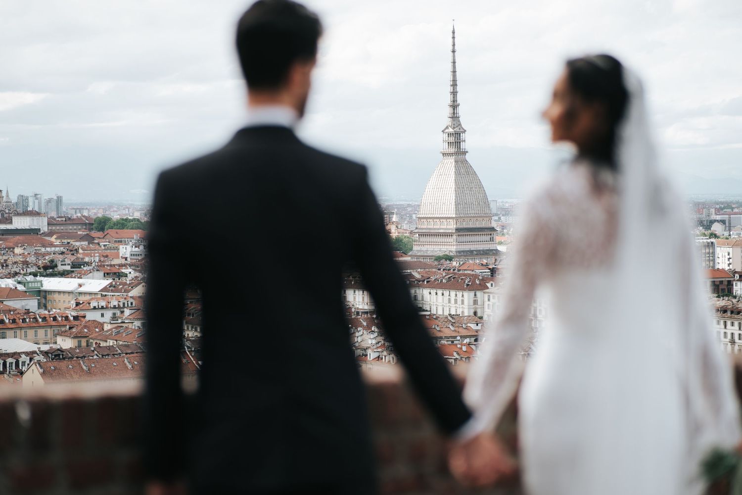 Bride and groom holding hands overlooking Turin cityscape with Mole Antonelliana on cloudy day.