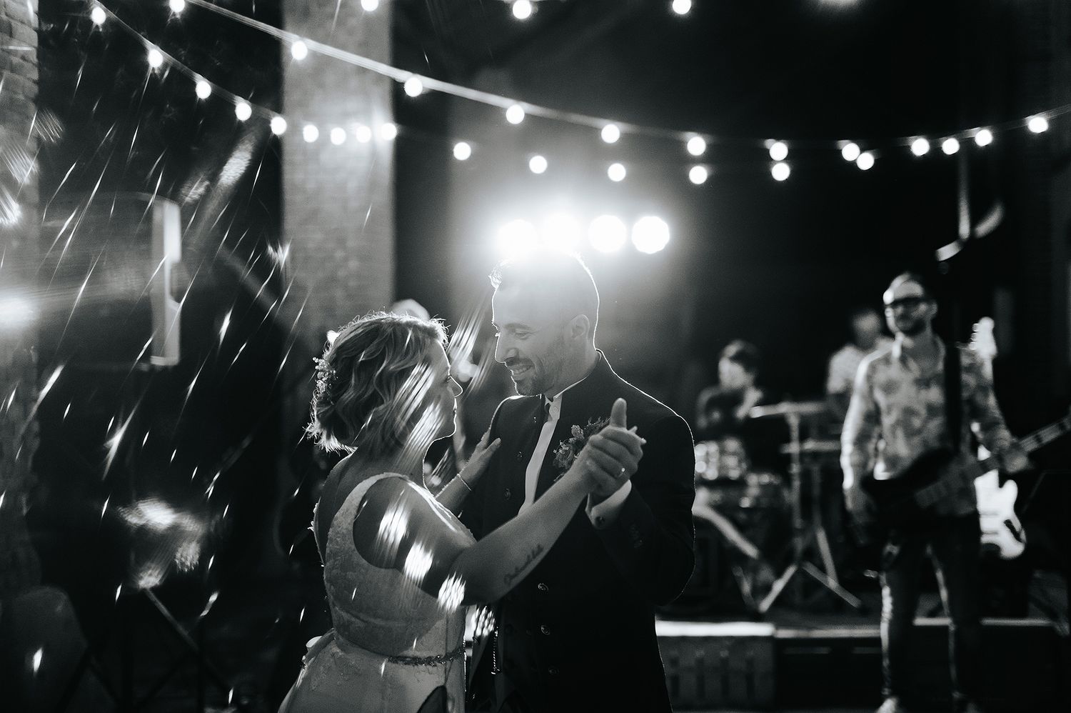 Black and white photo of bride and groom sharing first dance on stage with live band performing behind them.