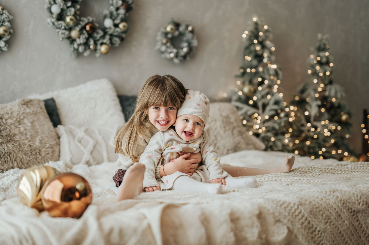 Smiling older sister embracing a joyful baby sibling in a cozy Christmas decorated bedroom.