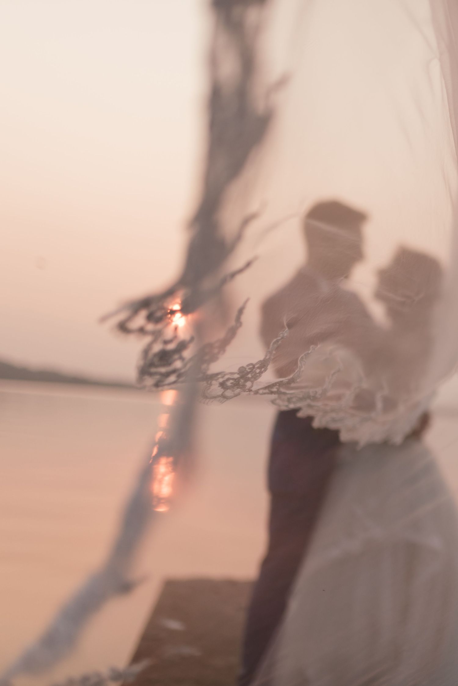 Dreamy soft-focus silhouette of bride and groom embracing through sheer lace veil at sunset beach wedding.