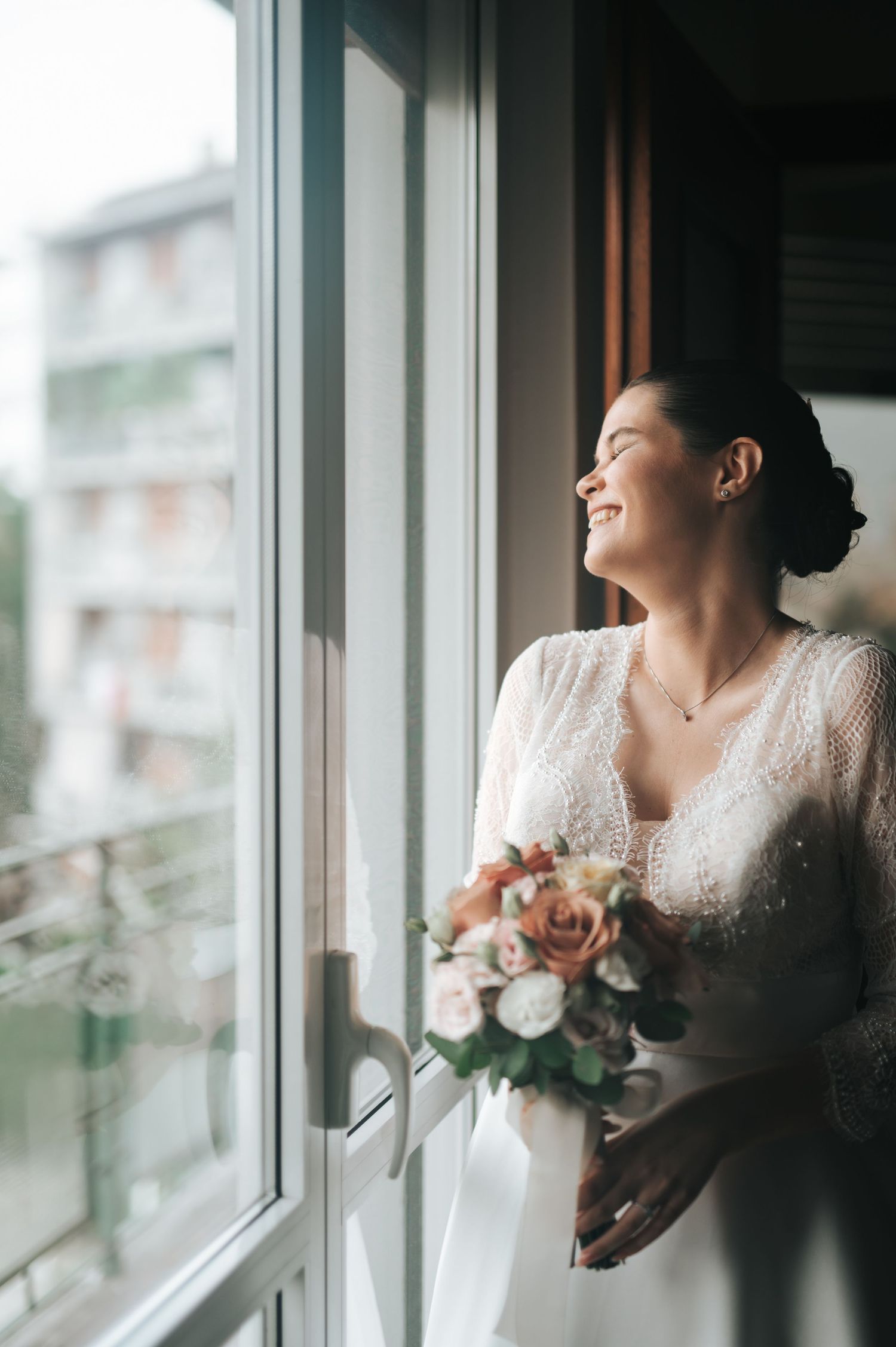 Radiant bride in lace gown holding pink rose bouquet, smiling by sunlit window on wedding day.