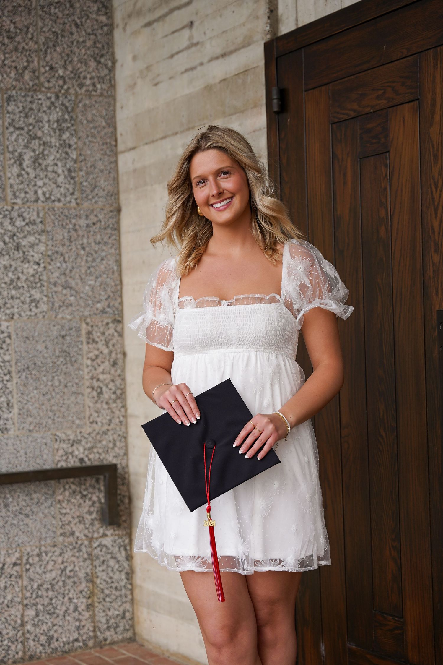 Graduate in white lace dress holds diploma with tassel against stone wall.