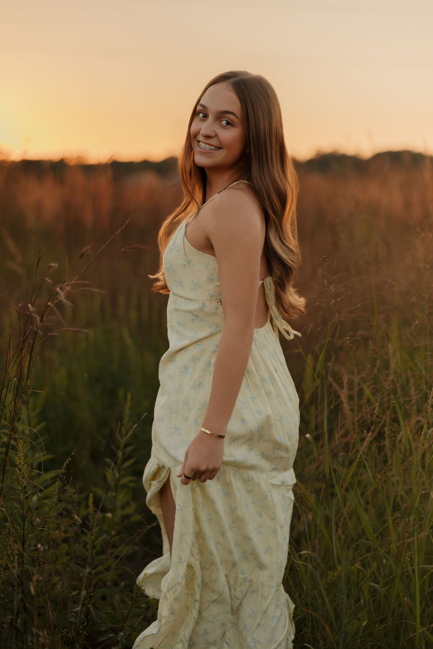 A woman in a flowing cream dress stands in a golden field at sunset, her long hair catching the warm evening light.