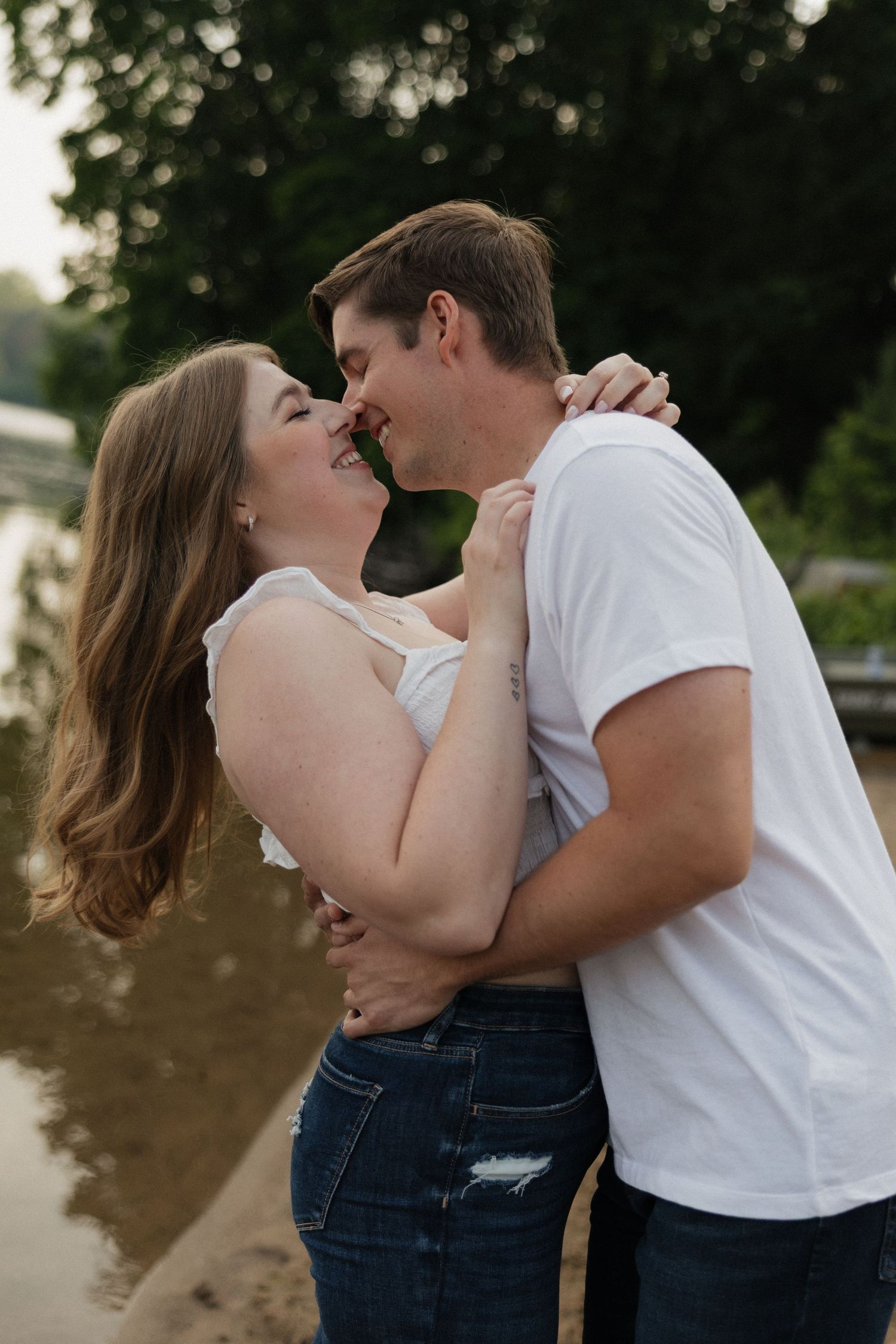 A romantic couple in casual outfits share an intimate moment by the water's edge during sunset.