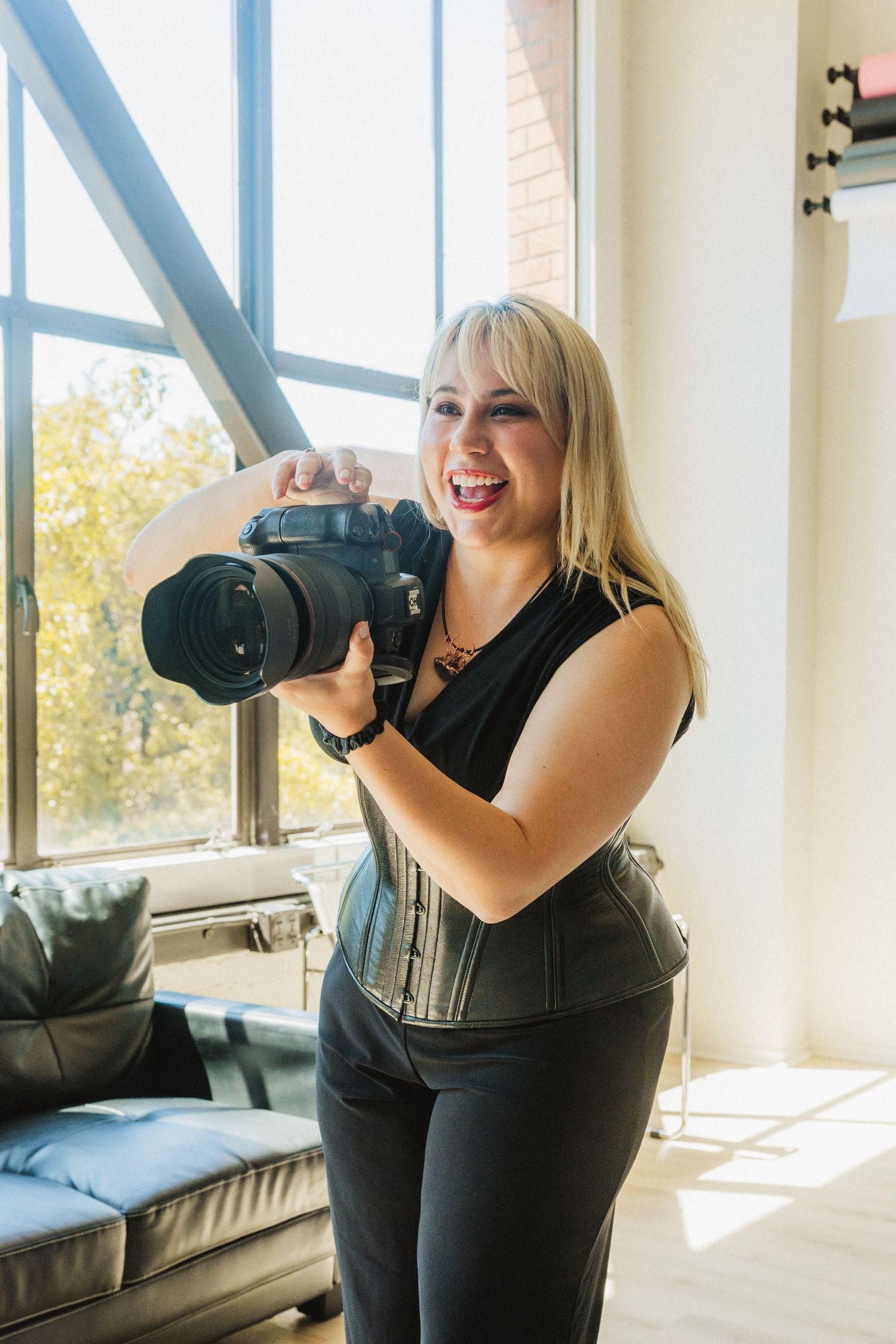 A photographer in black attire laughs while taking pictures with a professional camera in a bright indoor space.
