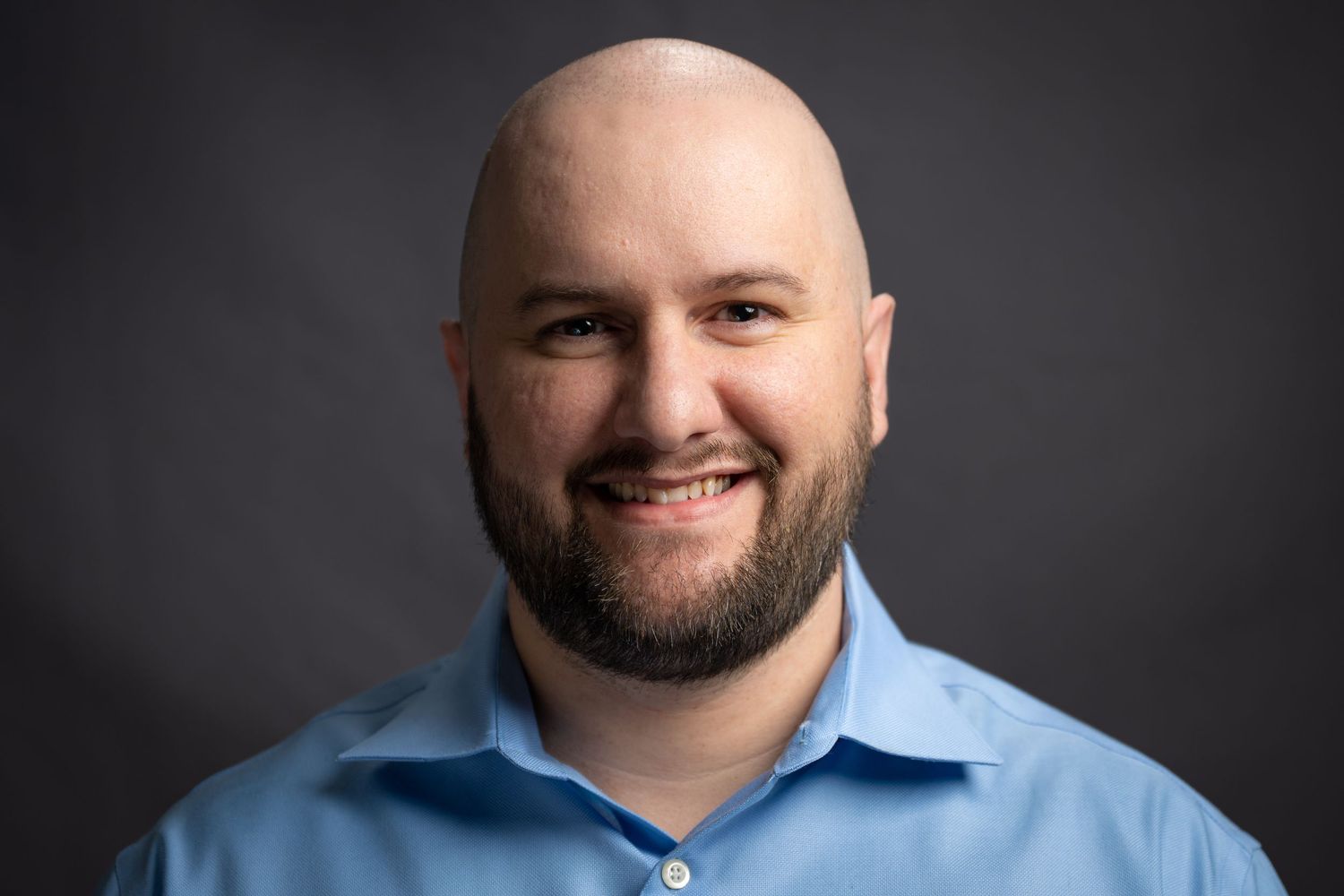 Professional headshot portraits of someone with facial hair wearing a blue collared shirt against a dark gray background.