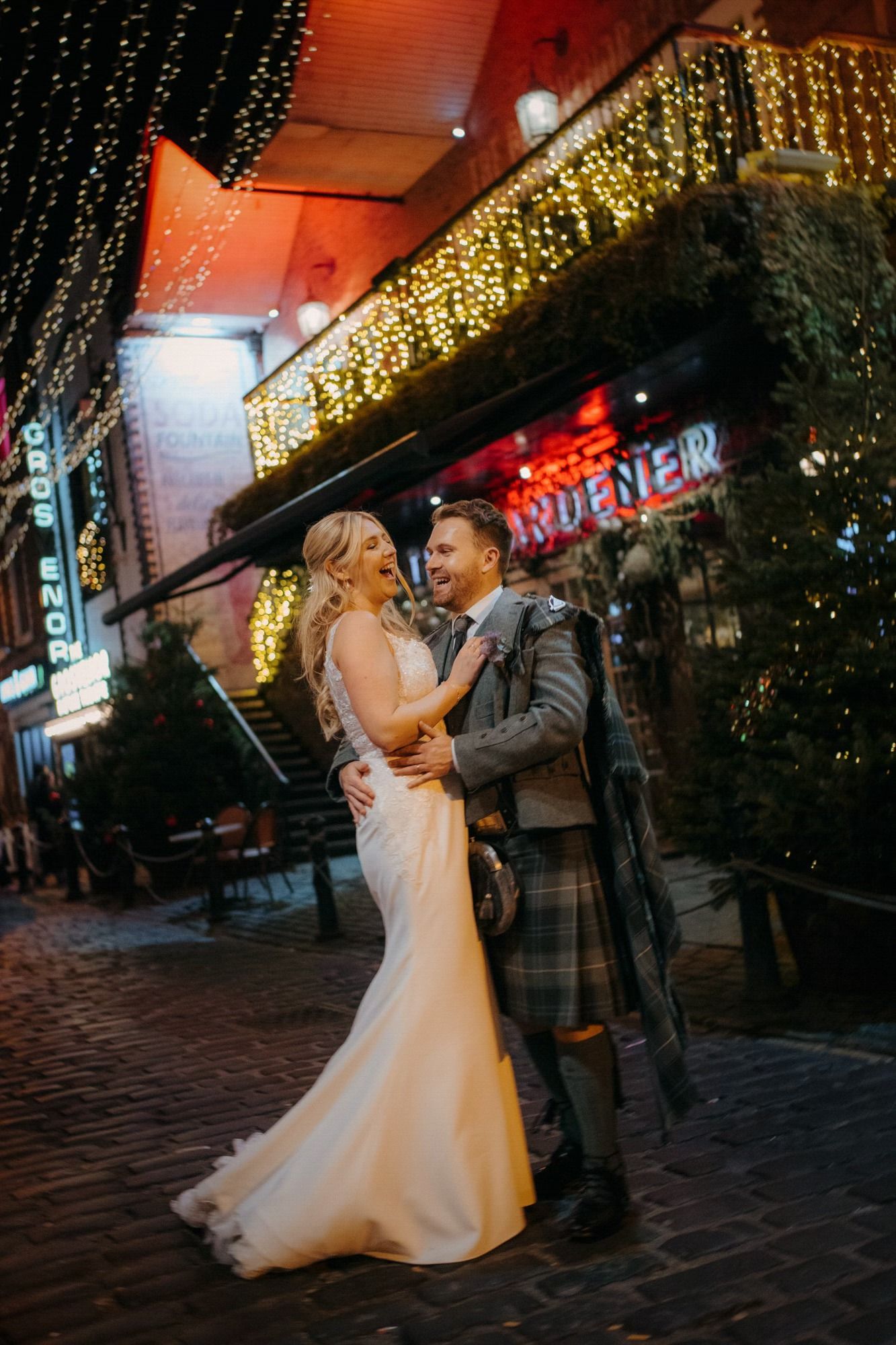 Newlywed couple shares romantic dance under twinkling lights in historic cobblestone street at night.