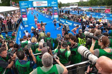 Photographers with professional cameras gather at a sporting event behind blue barriers to capture the action.