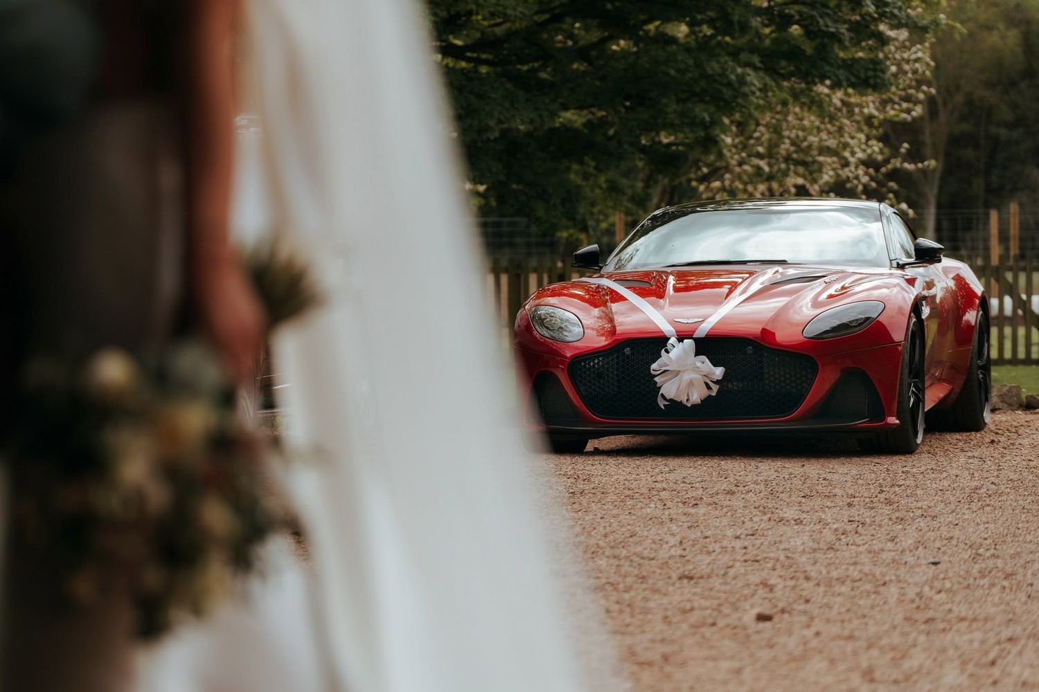 Red Aston Martin wedding car decorated with white ribbon bow on a gravel driveway at an elegant venue.
