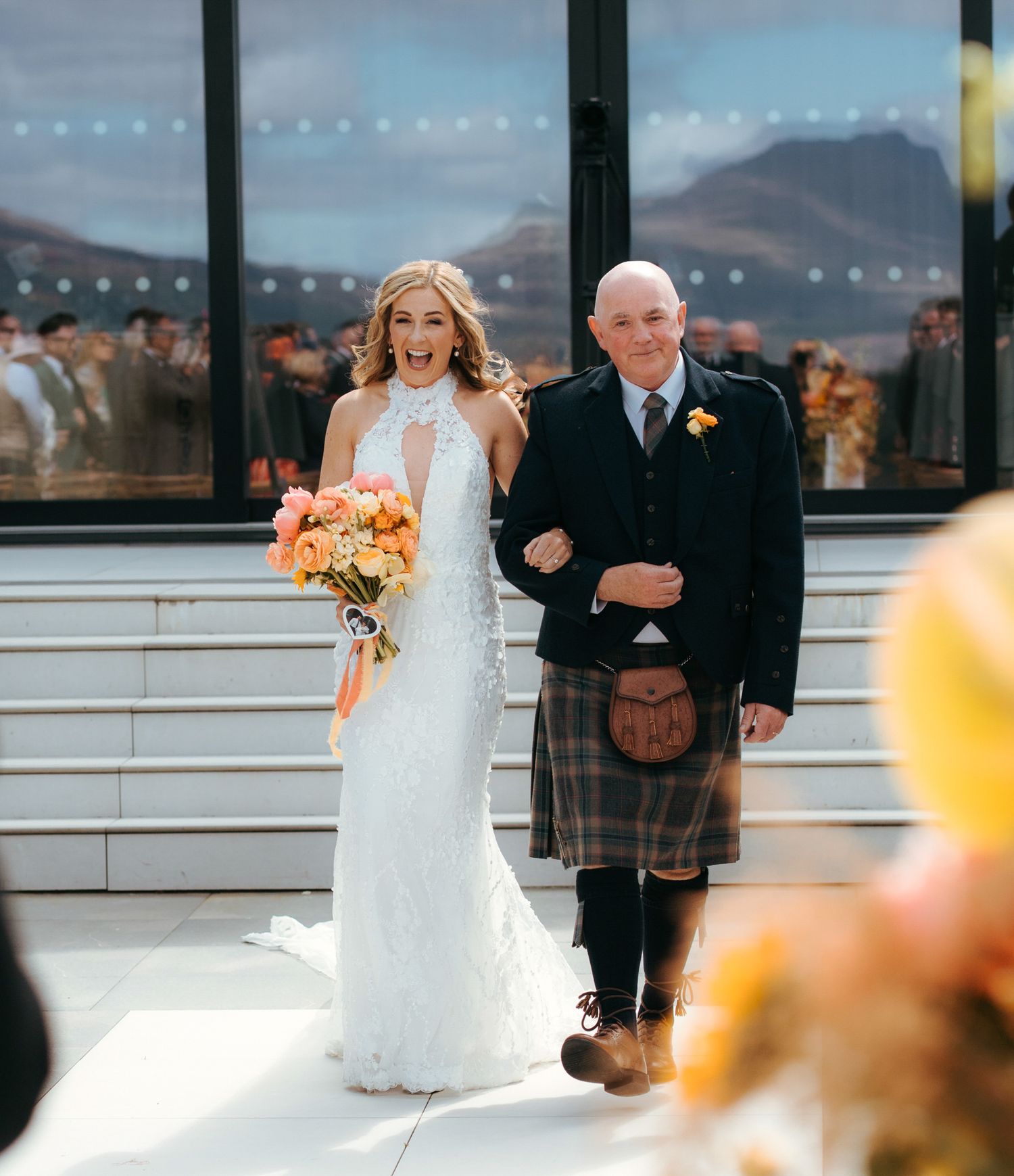 A bride in a white lace gown walks with a gentleman in traditional Scottish kilt attire on a wedding day.