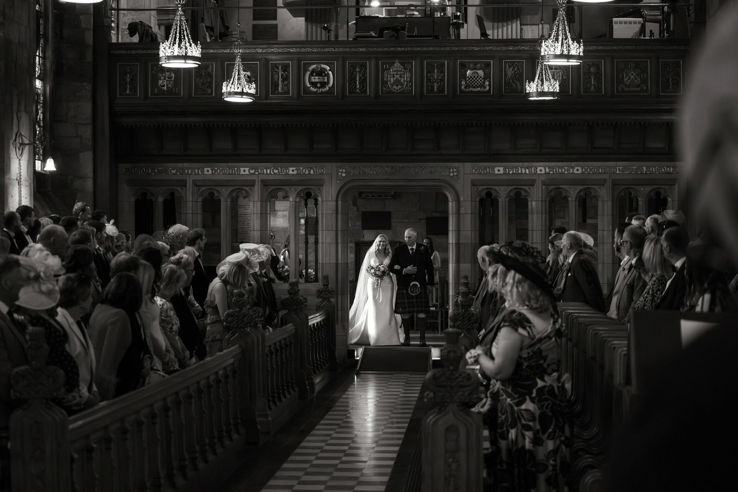 Black and white wedding ceremony photo inside an ornate church with guests seated in wooden pews as couple stands at altar.