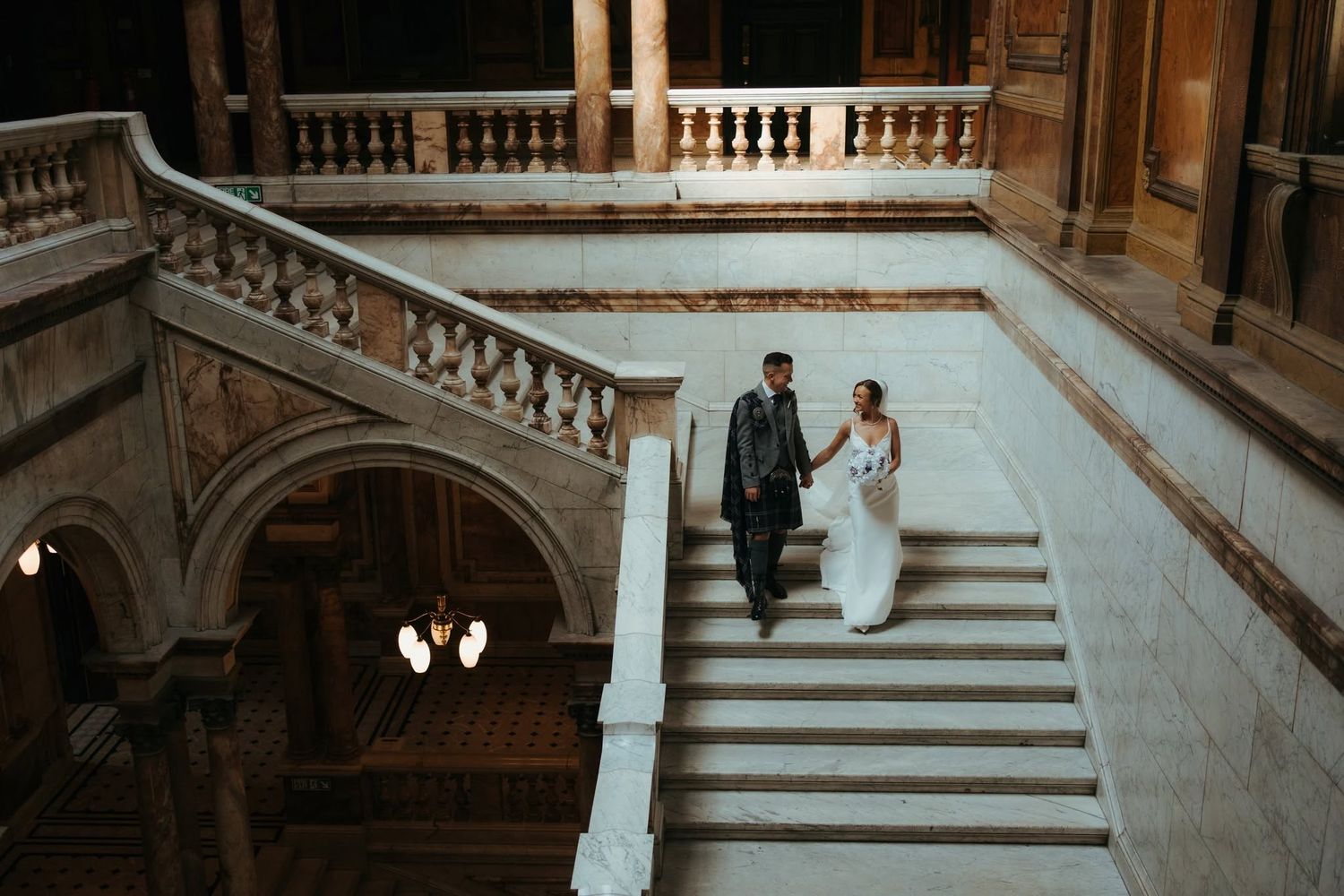 Bride and groom walking up grand marble staircase in elegant historic building during wedding photo session.