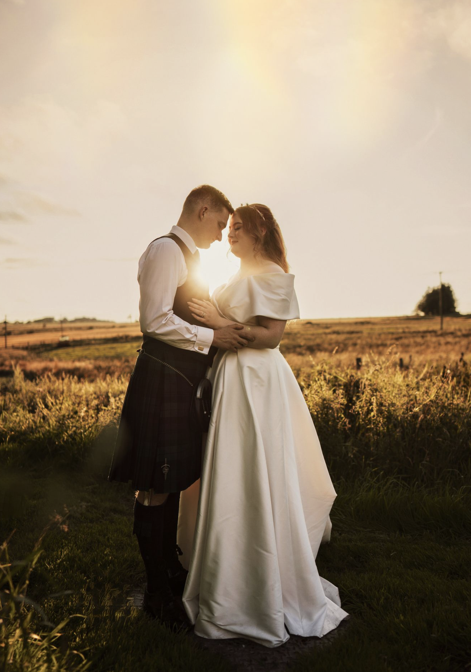 A couple in wedding attire embraces in a golden field at sunset, creating a romantic silhouette against the glowing sky.
