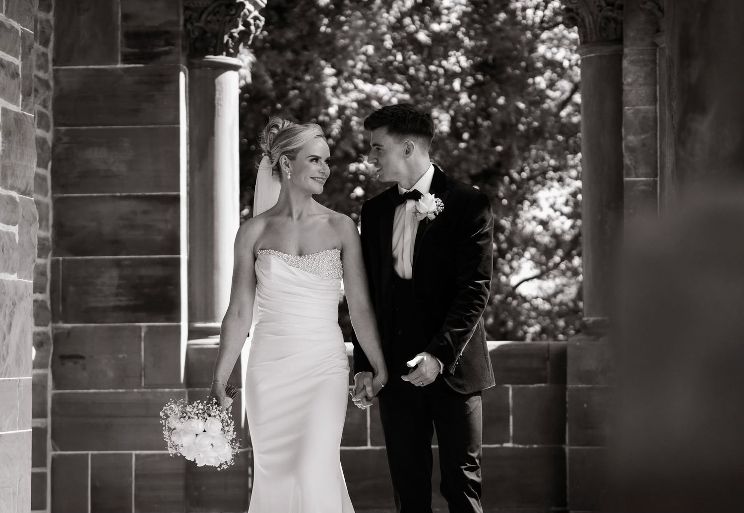 Black and white wedding photo of bride in strapless gown and groom in tuxedo holding hands near stone archway.