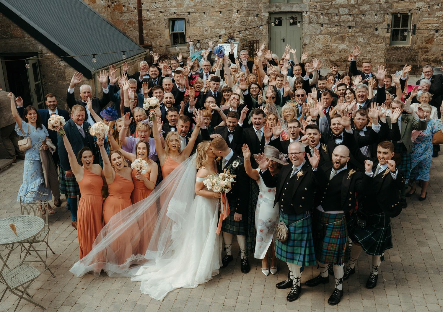 Wedding guests gather in a courtyard with brick walls as the couple poses for a panoramic group photo.