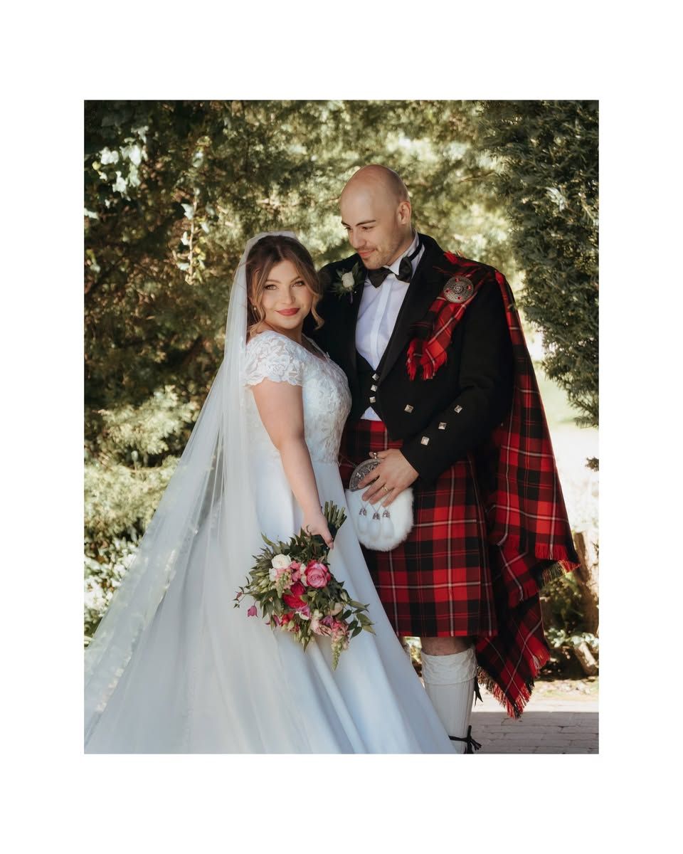 Bride in white gown holding pink bouquet stands with groom in traditional red Scottish kilt attire outdoors.