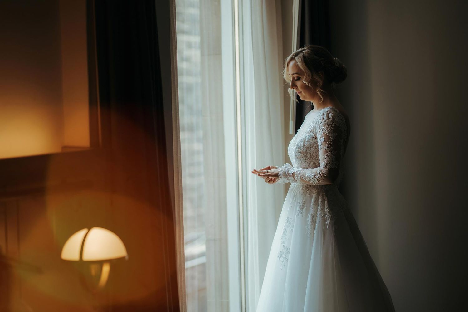 A radiant figure in a white lace wedding gown stands by a sunlit window, creating a romantic silhouette.