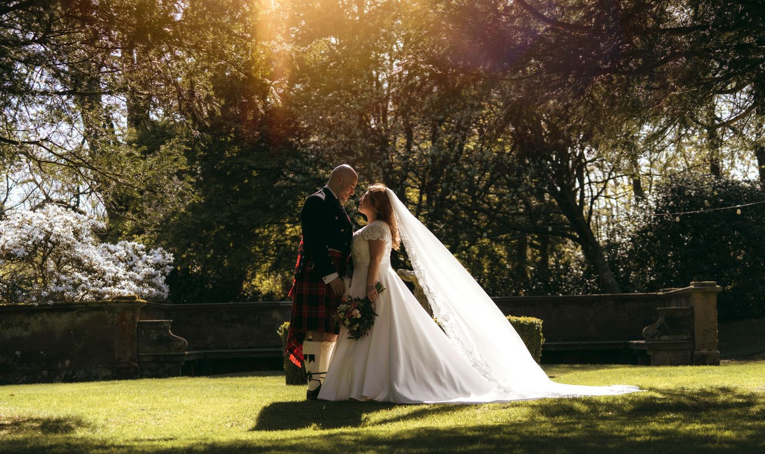Bride and groom share a romantic moment in a sunlit garden, groom wearing a traditional Scottish kilt.