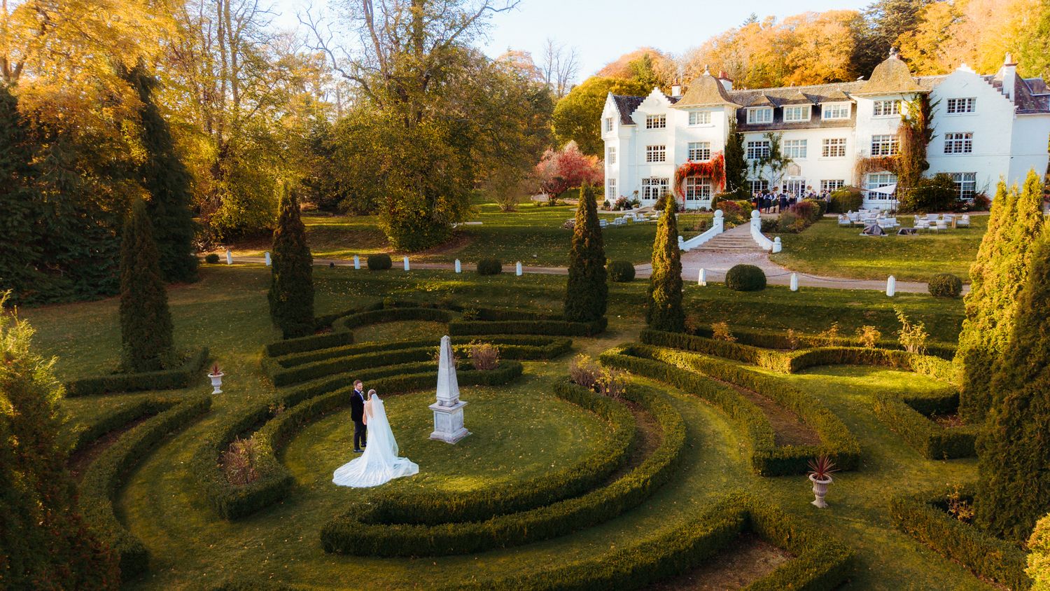 A scenic autumn garden with ornate hedgerow mazes and pathways surrounded by golden fall foliage and white buildings.