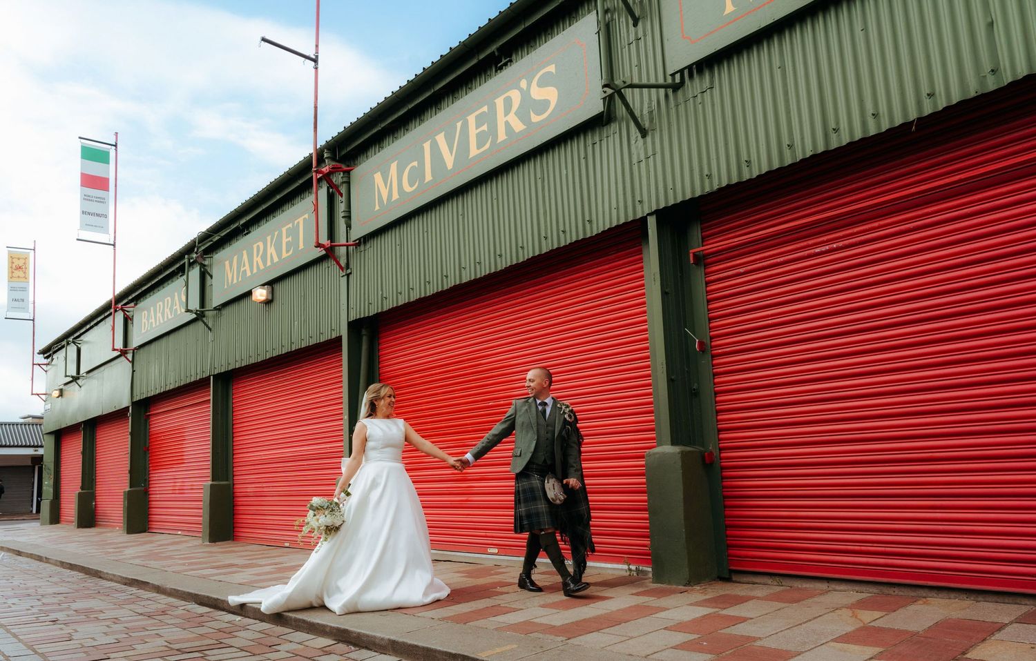 Bride and groom holding hands outside McIver's building with red roller doors in Scotland.