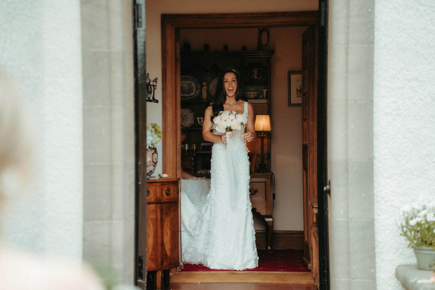 A bride in a white wedding gown stands in a doorway holding a bouquet while getting ready for her wedding ceremony.