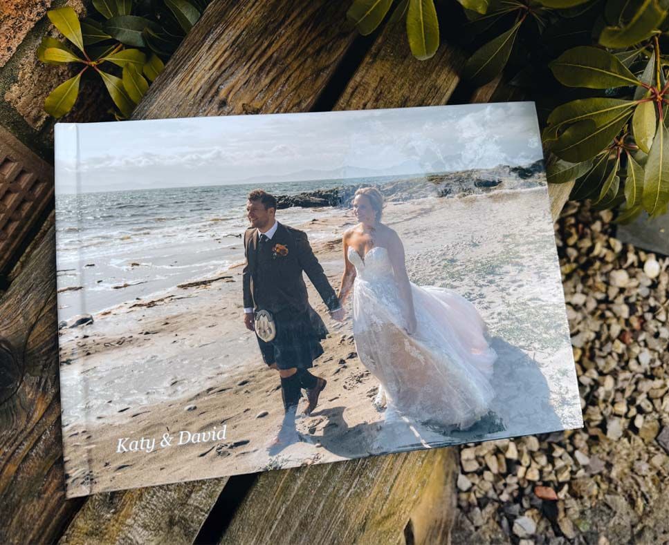 A printed wedding photograph lying on a wooden surface with green foliage, showing a couple walking along a beach.