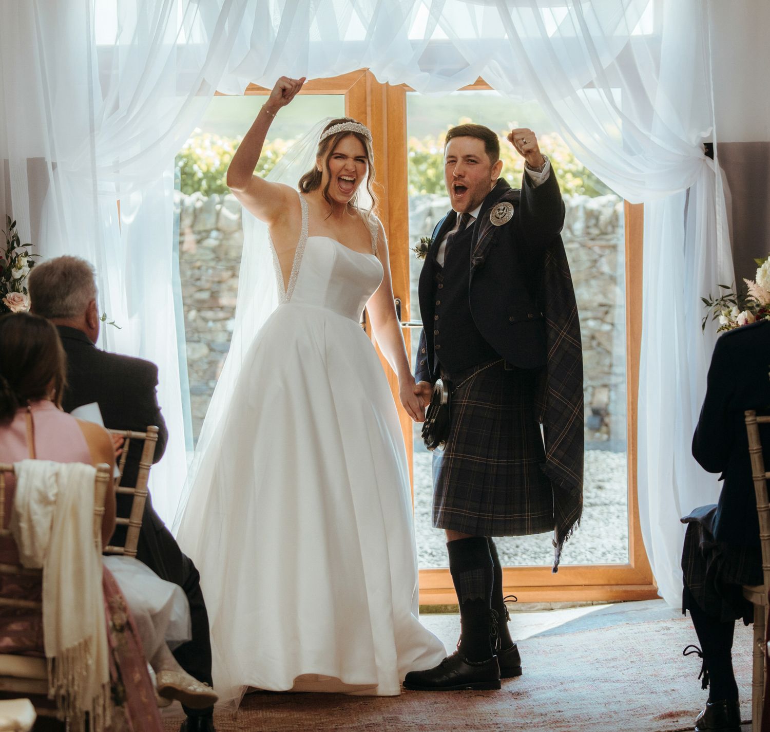 Joyful newlyweds celebrate at wedding ceremony, bride in white gown and groom in traditional Scottish tartan kilt, raising fists.