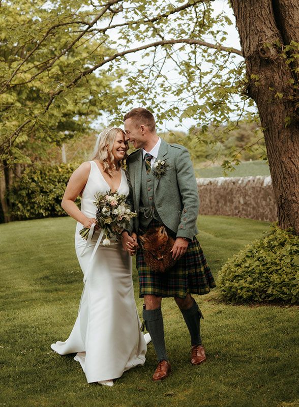 A bride and groom share a romantic moment outdoors, she in a white gown with bouquet, he in a green tartan kilt.
