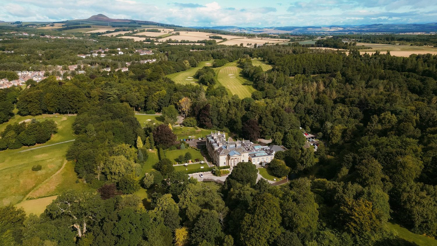 Aerial view of a grand country estate surrounded by lush green woodland with rolling hills and mountains in the background.
