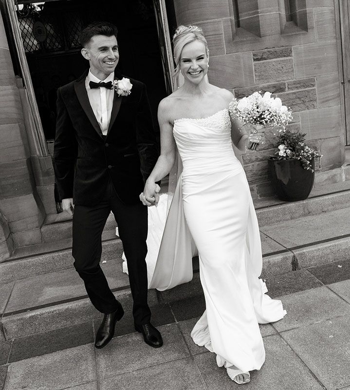 Black and white photo of smiling bride and groom holding hands outside church, bride in strapless gown holding bouquet.