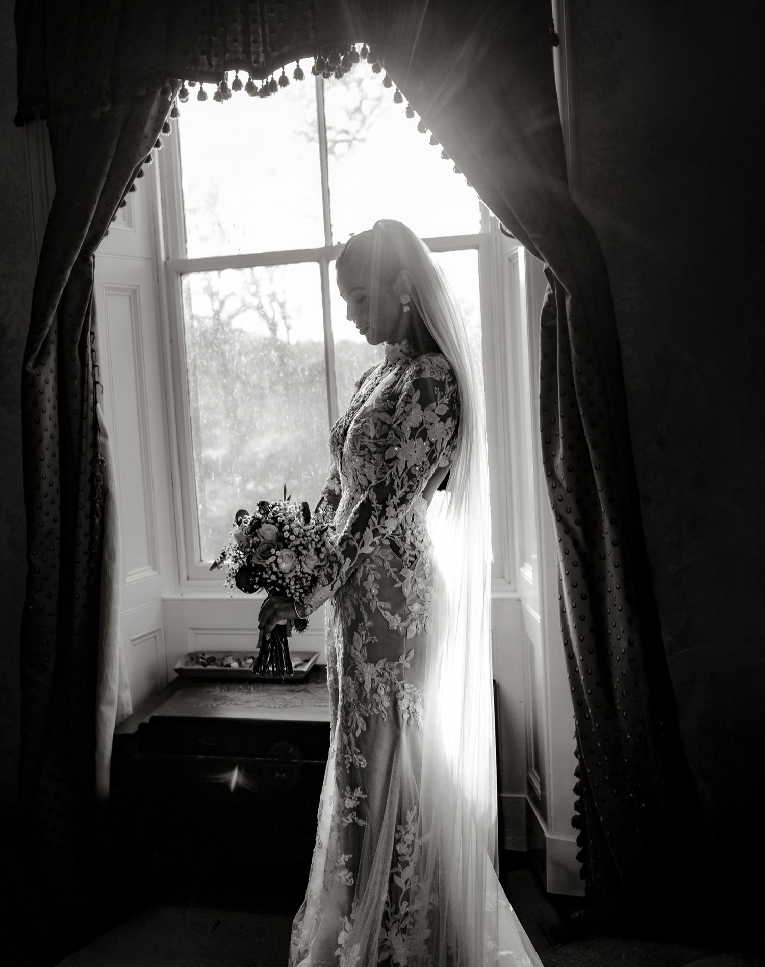 A silhouetted figure in a flowing wedding dress stands by a window in dramatic black and white photography.