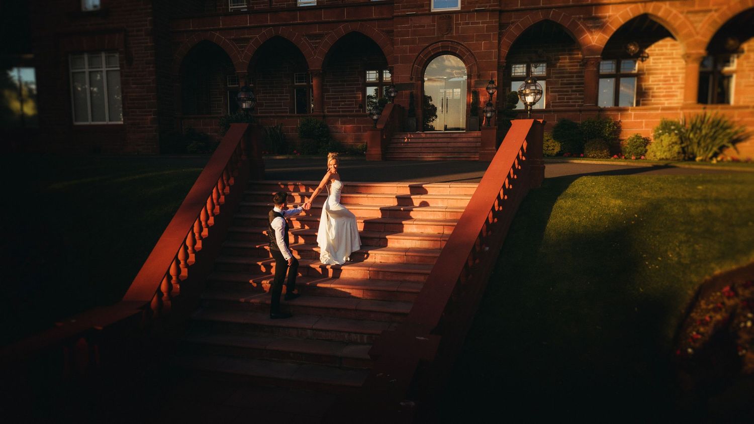 Bride in white dress descends sunlit brick staircase of historic stone building during golden hour.