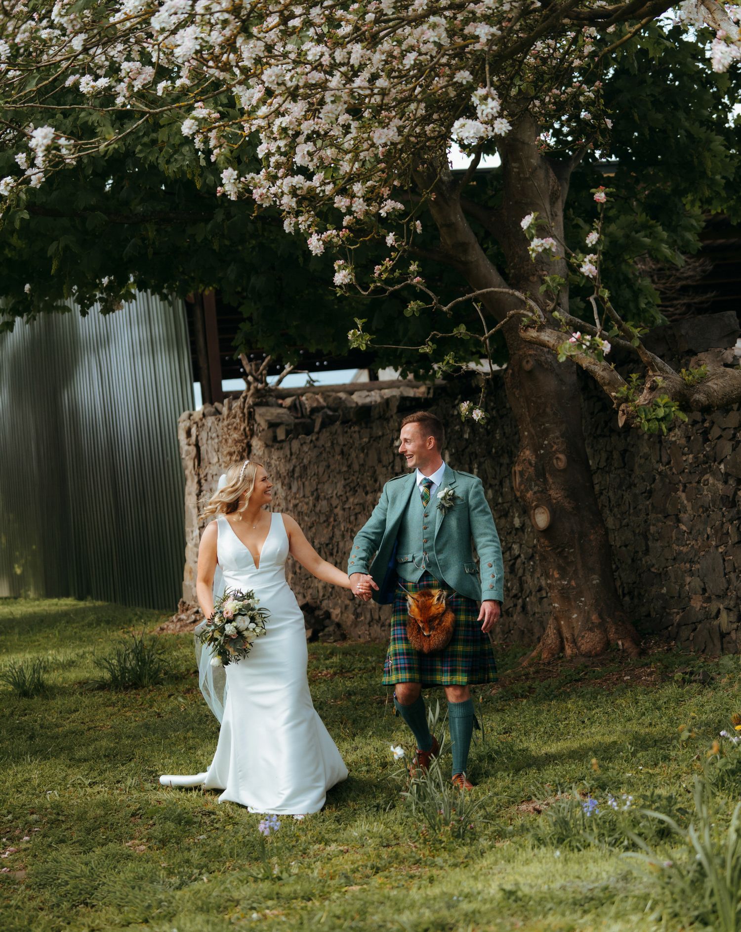 A bride and groom walk hand in hand under a blossoming tree, the groom wearing a traditional Scottish tartan kilt.