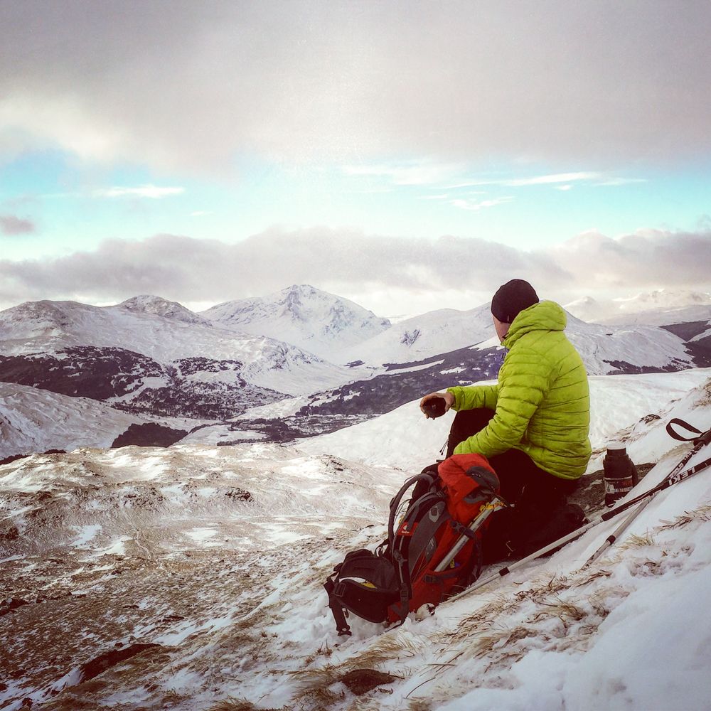 A mountaineer in a bright green jacket sits on a snowy peak, gazing at a dramatic winter mountain landscape under cloudy skies.