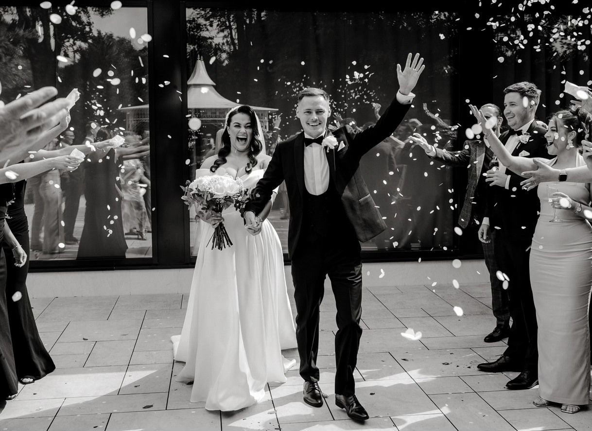 Newlywed couple walks through a shower of white flower petals on their wedding day while guests celebrate.