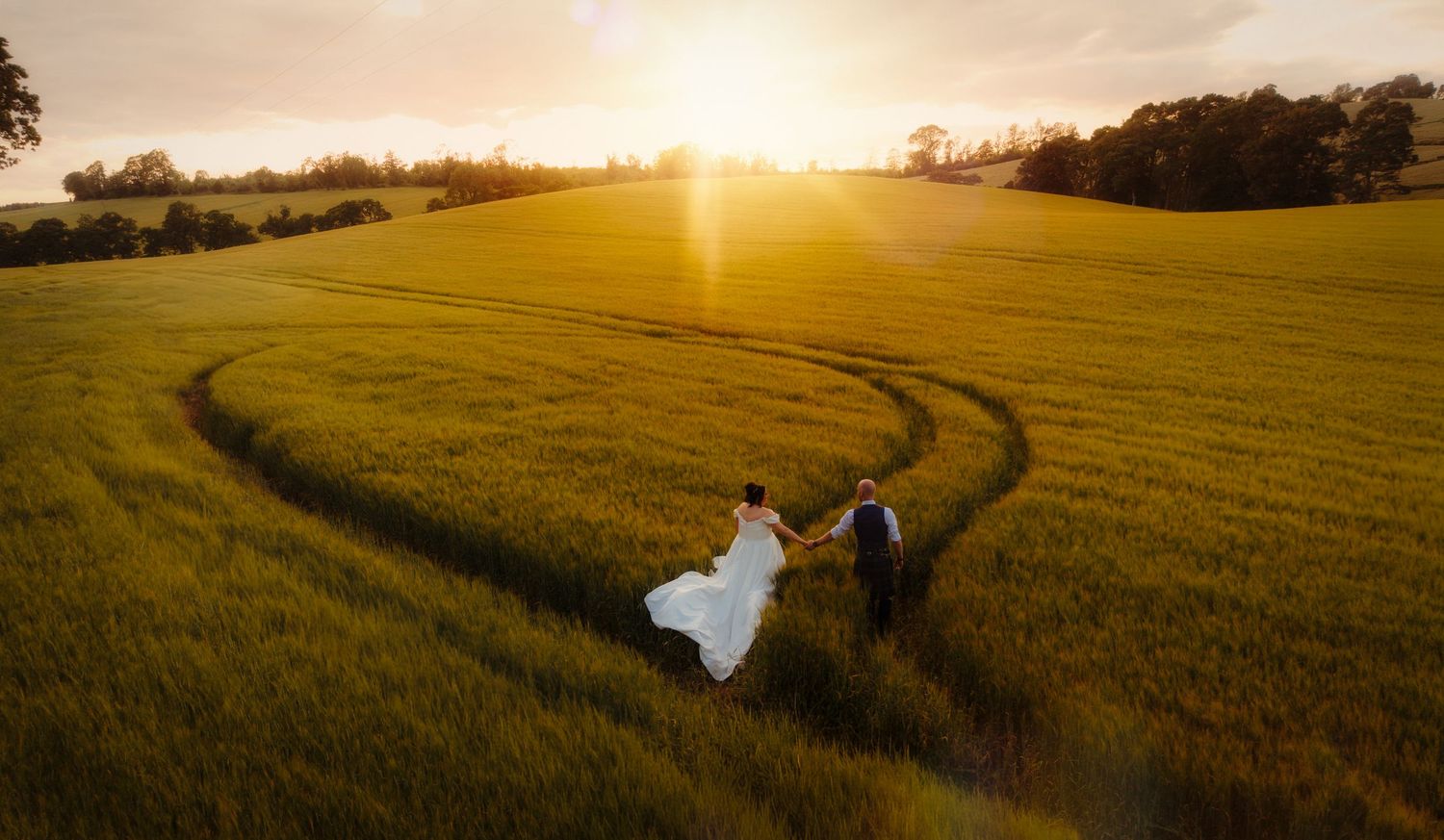 Aerial view of bride and groom walking through lush green field at golden sunset, wedding photography.