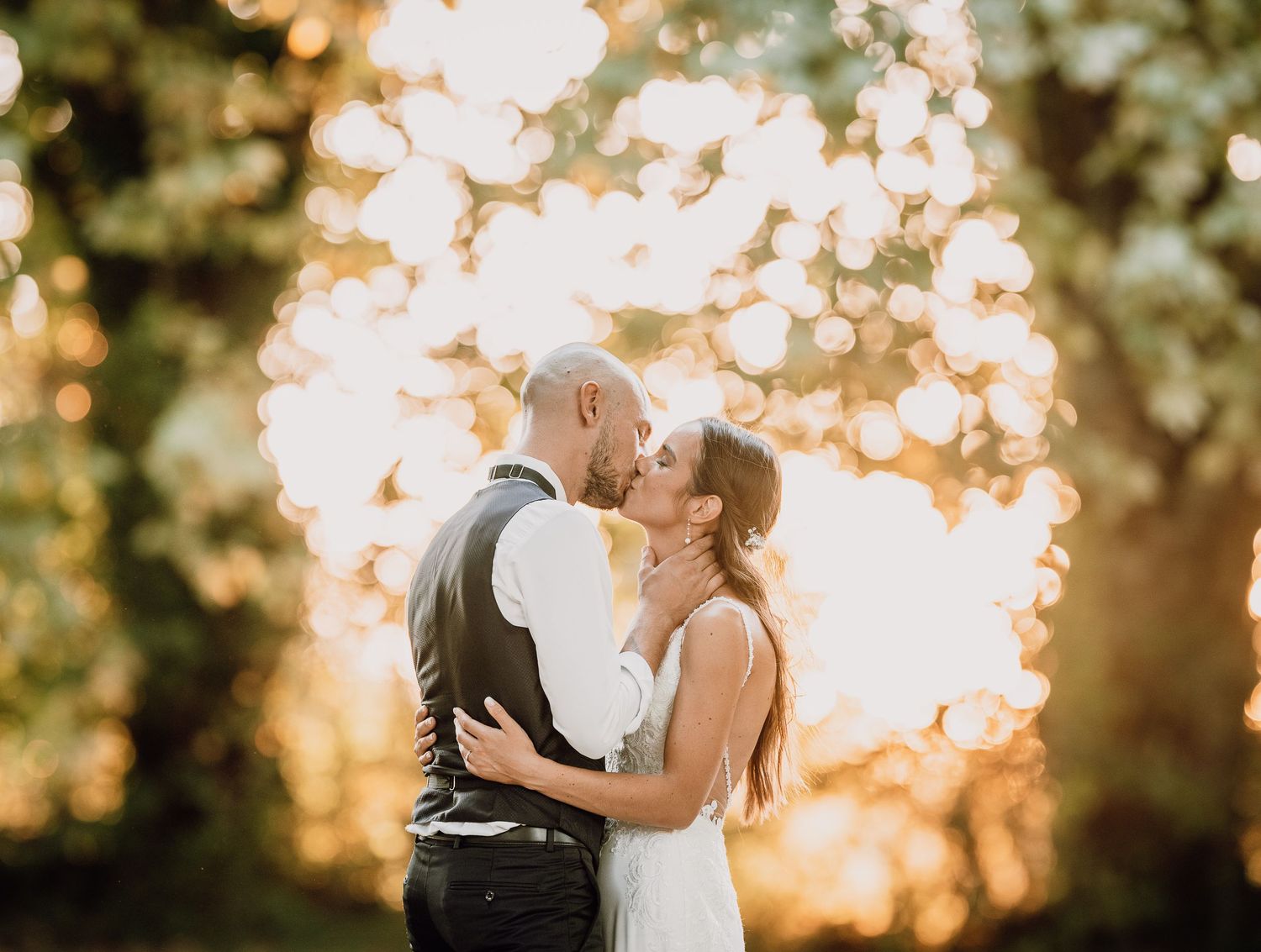 A couple shares a romantic kiss at sunset with glowing golden bokeh lights and trees in the background.