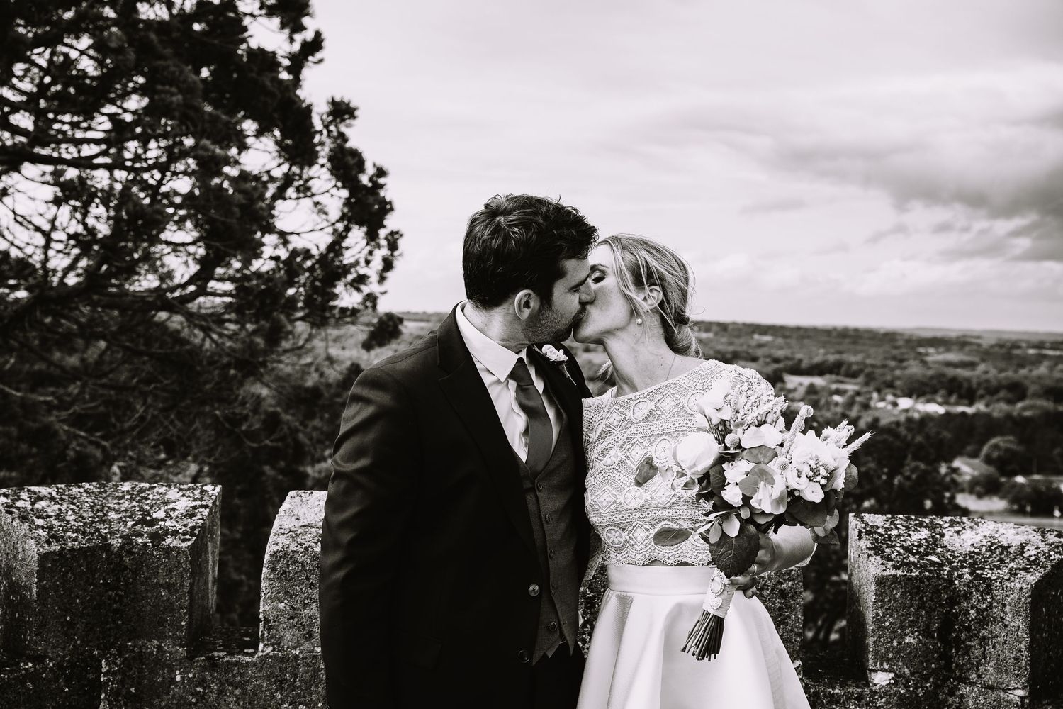 A romantic black and white wedding photo of a couple sharing a tender moment on a hilltop overlooking a scenic landscape.