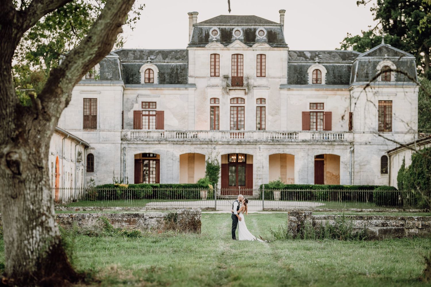 A romantic couple stands in front of a historic white mansion with ornate architecture and lush green grounds.