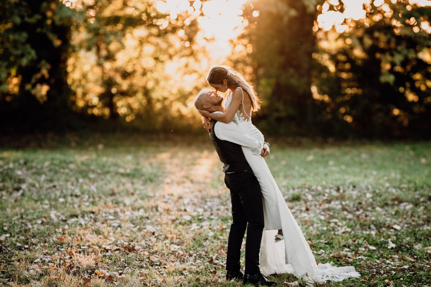 A romantic couple embraces in a sunlit autumn garden with fallen leaves scattered on the ground during golden hour.