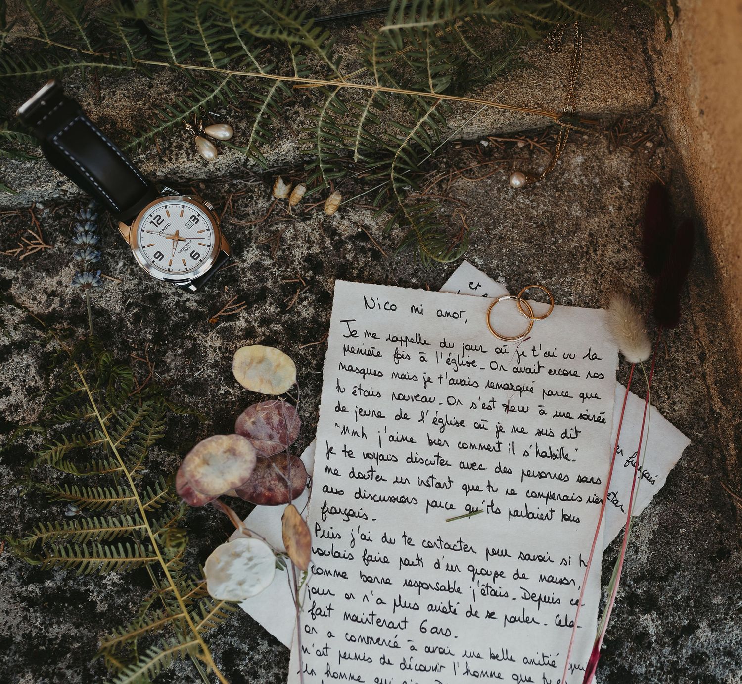 Détails de mariage : lettre manuscrite, alliances, montre et coquillages sur un lit de fougères