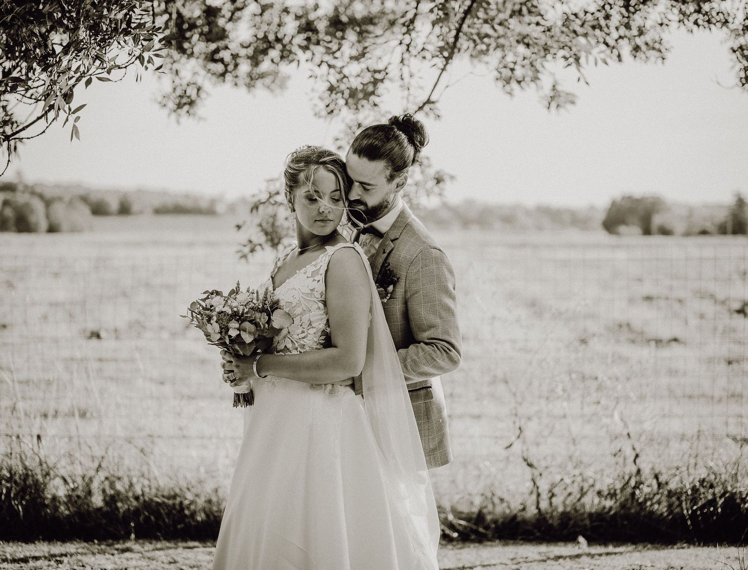 A black and white wedding photo of a couple embracing in a sun-lit field with the bride holding a floral bouquet.