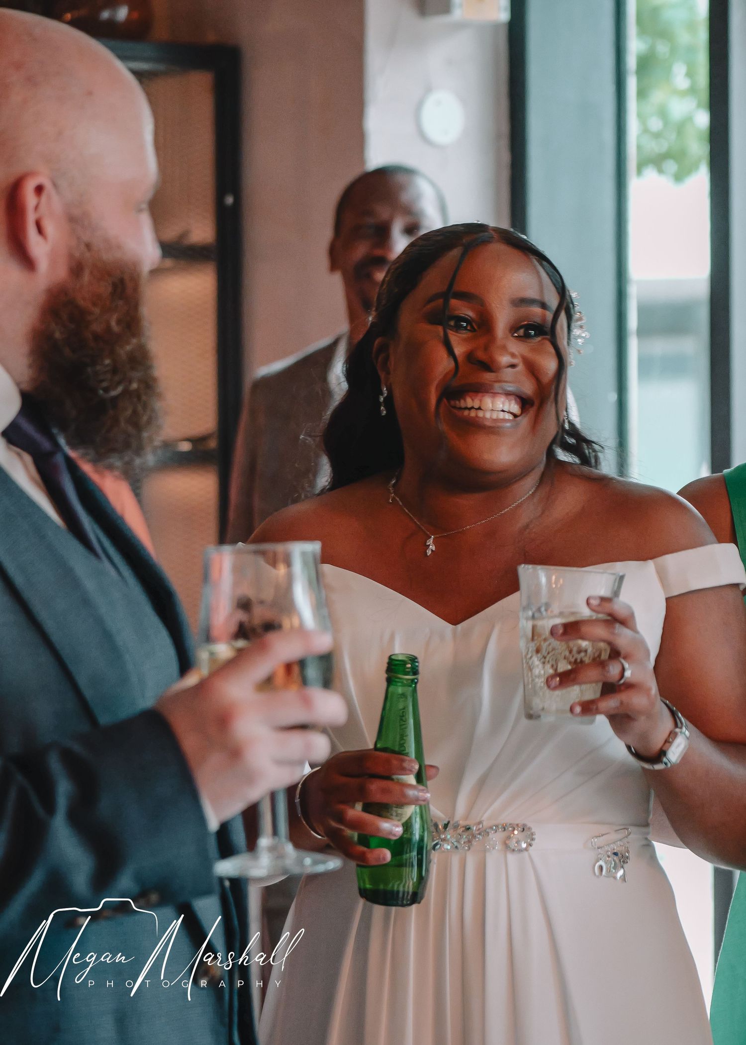 Joyful bride in white off-shoulder gown laughing and holding drinks with guests at wedding reception.