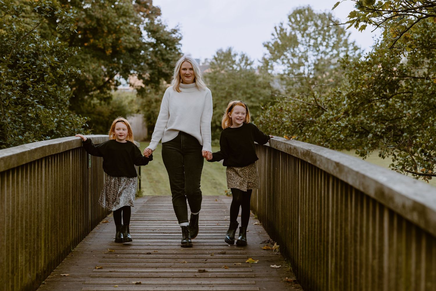 Family Photoshoot - Three people holding hands while walking together on a wooden boardwalk surrounded by green trees on an autumn day.