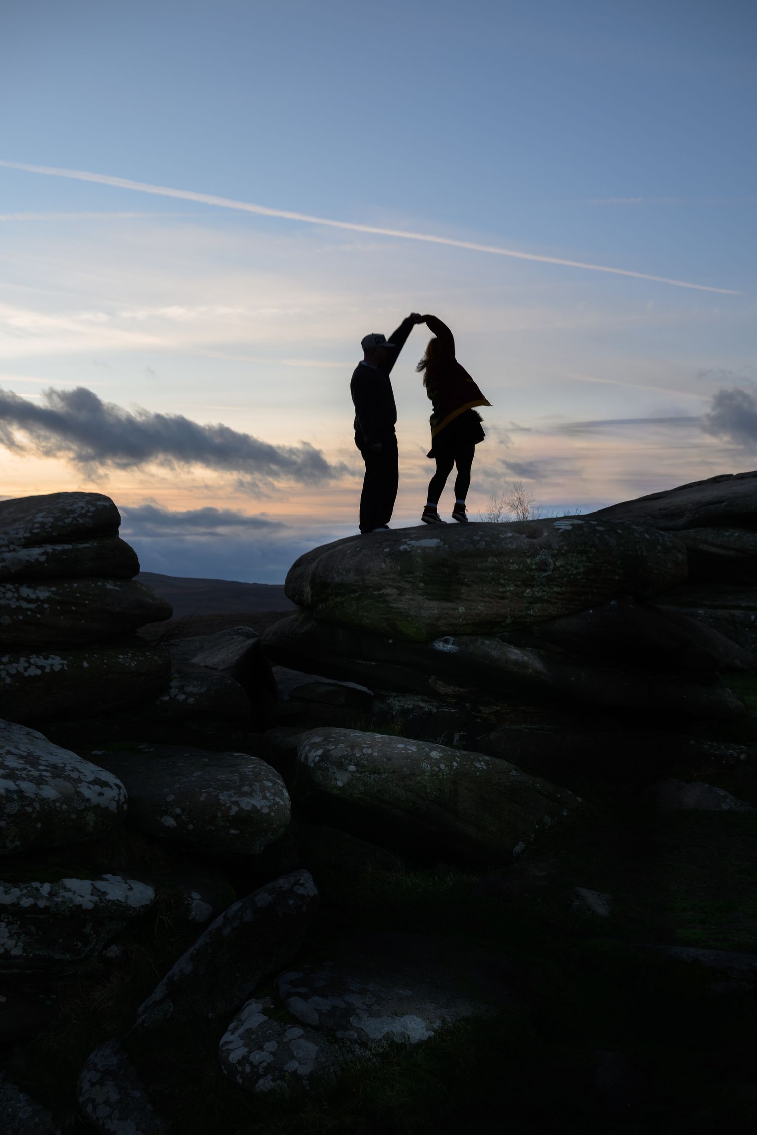 Silhouette of a couple dancing on rocky hilltop at sunset with dramatic cloudy sky backdrop.