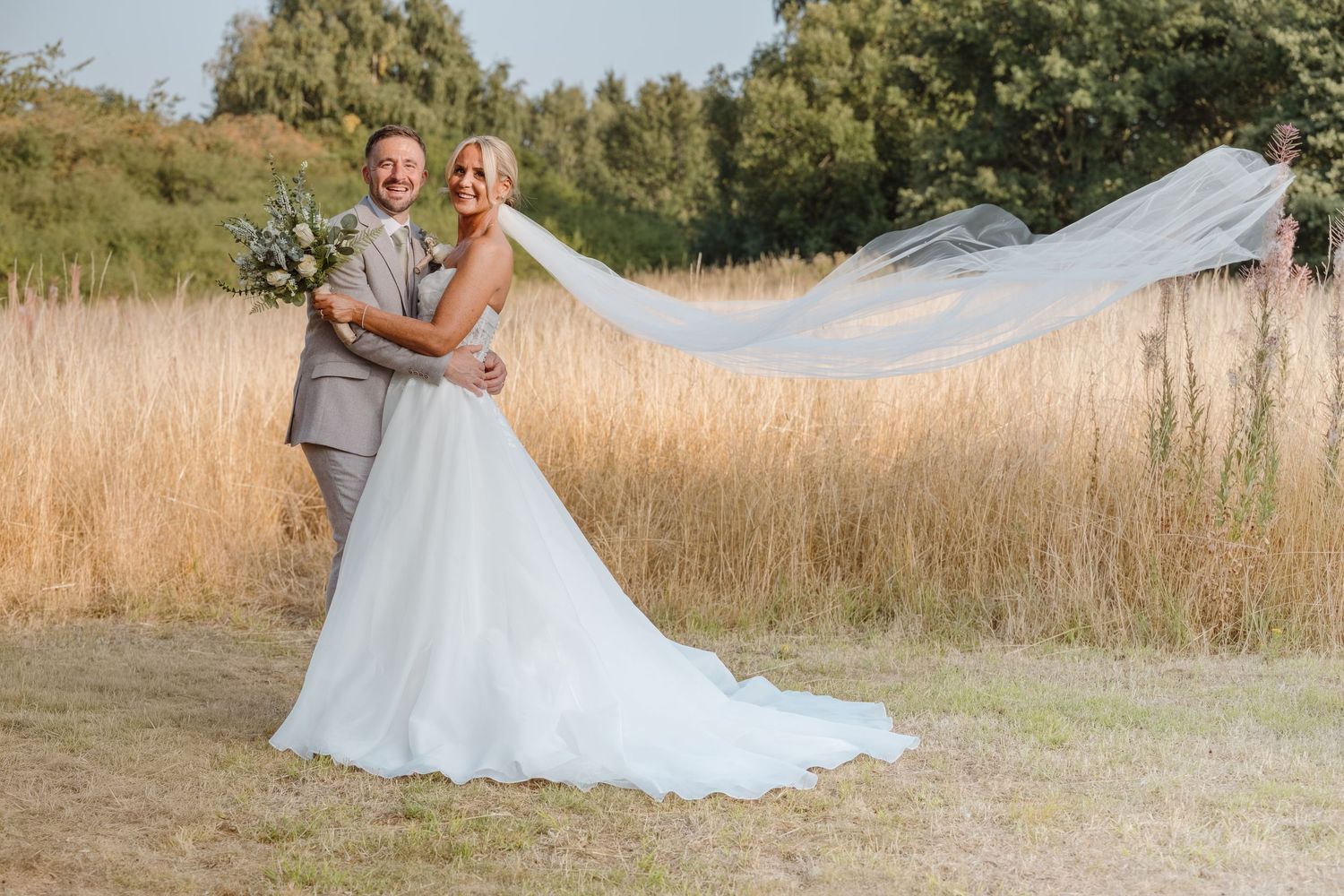 Bride and groom embrace in golden field, her veil flowing in the wind, wearing white gown and gray suit.