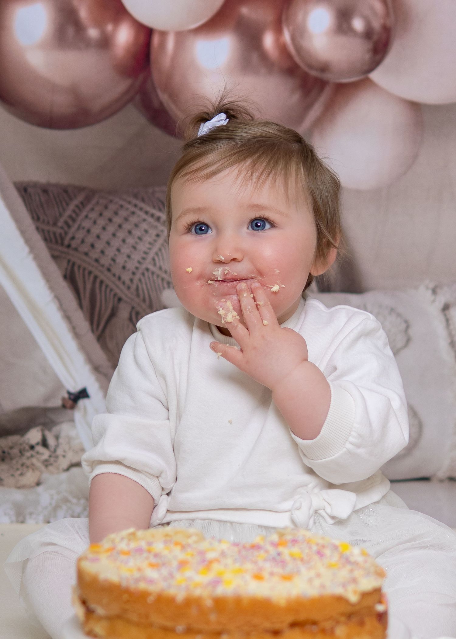 A baby in white clothing reaches for first birthday cake surrounded by pink balloons. Cake smash photoshoot